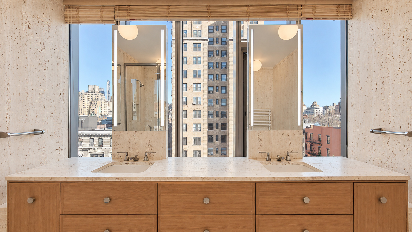 Pale double vanity countertop beneath two full mirrors and tall windows, surrounded by travertine walls