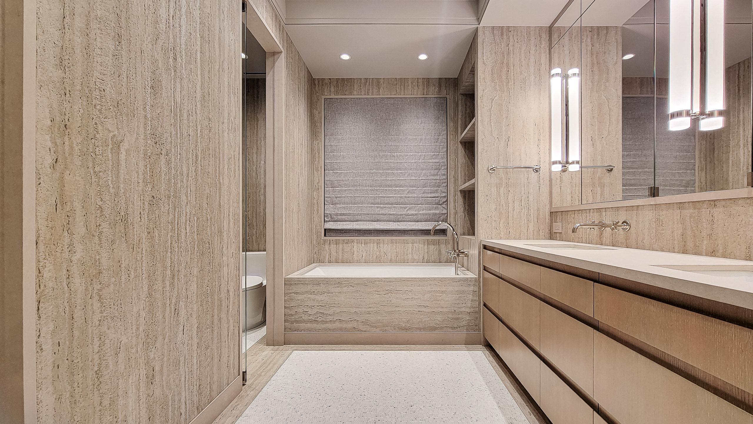 Warm modern bathroom with beige slab walls, a wood vanity, and a built-in bathtub.